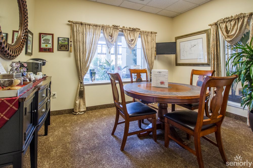 A cozy dining room with a wooden table and chairs, complemented by curtains and a carpeted floor.
