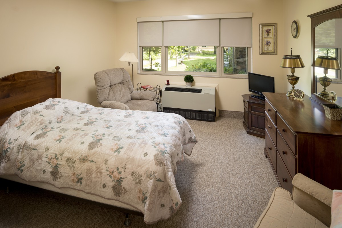 A cozy bedroom with a bed, armchair, and a small television, featuring a window with a view outside.