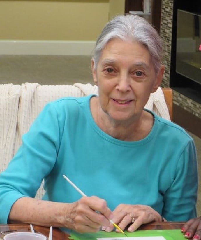An elderly woman in a blue shirt is sitting indoors and writing.