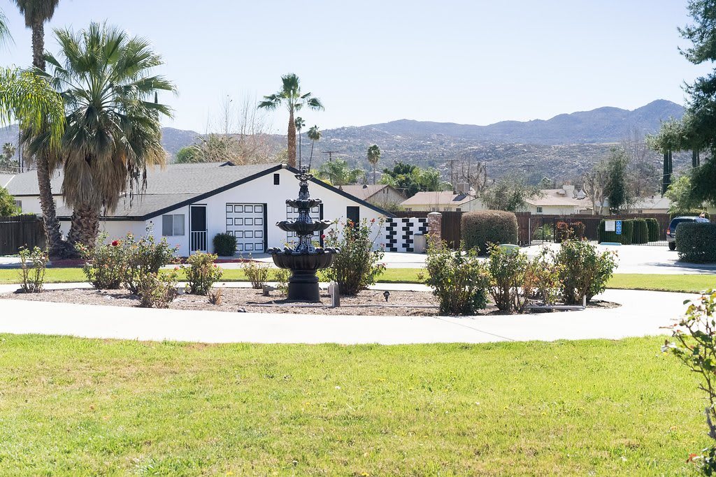 A scenic view of a house with a central fountain, surrounded by palm trees and mountains in the background.