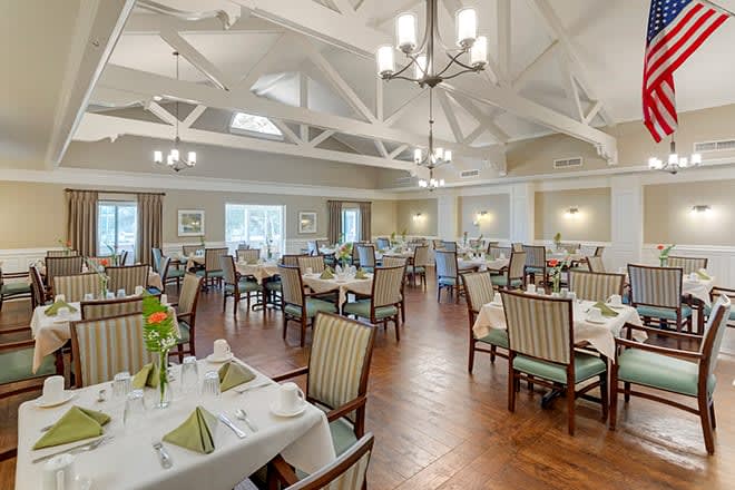 An elegantly set dining hall with multiple tables and chairs, featuring chandeliers and natural lighting.