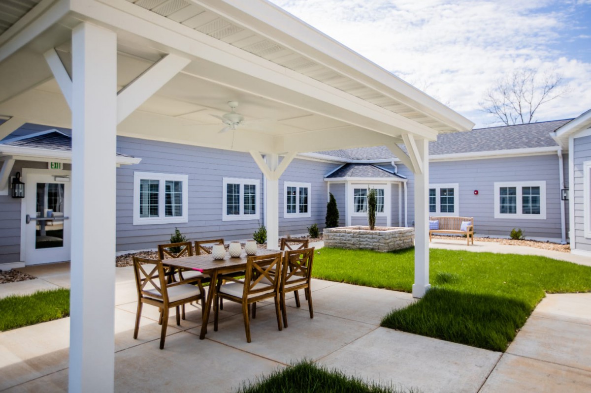 A covered patio with a dining table set in the backyard of a house, featuring a well-maintained lawn and landscaping.