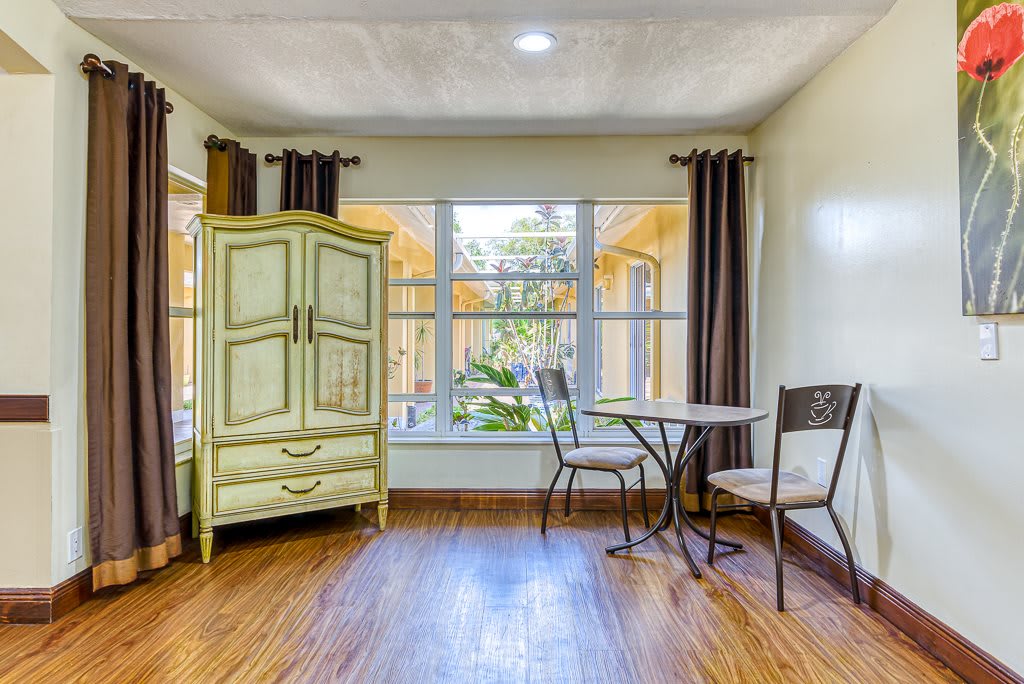 A cozy interior room with a small table and chairs, a vintage cabinet, wooden flooring, and large windows letting in natural light.