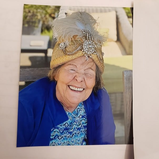 An elderly woman wearing a decorative hat, smiling brightly while seated outdoors.