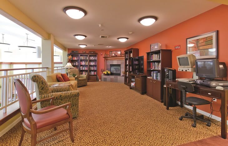 A cozy living room with bookshelves, comfortable seating, and a desk with a computer, illuminated by warm lighting.