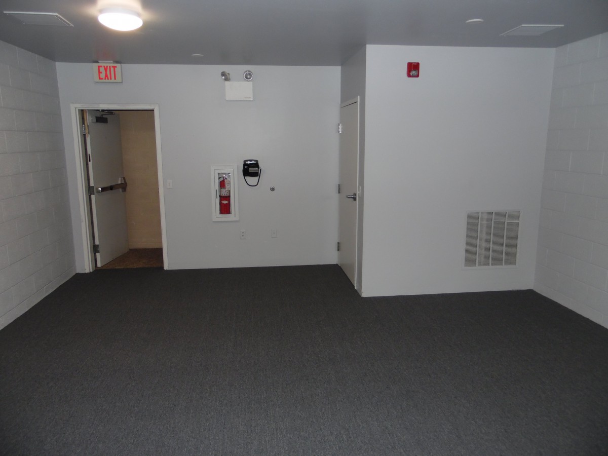 An empty interior space with a carpeted floor, white walls, an exit door, and fire safety equipment.
