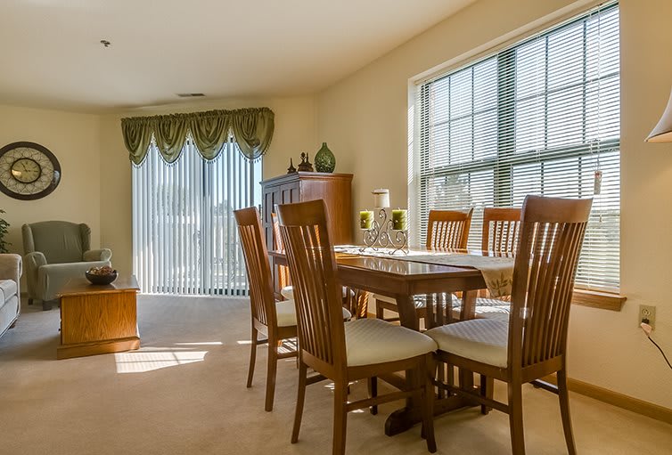 A well-lit dining room with a wooden table and chairs, featuring large windows allowing in natural light.