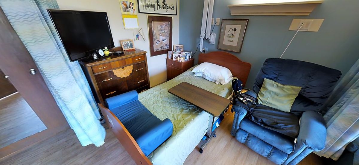 A cozy bedroom featuring a hospital bed, wheelchair, and wooden furniture, adorned with framed artwork on the walls.