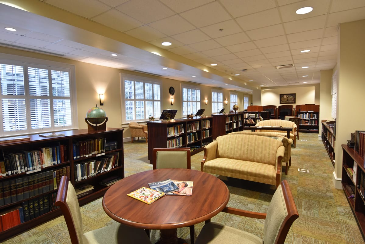 A cozy library interior featuring bookshelves, comfortable seating, and large windows.