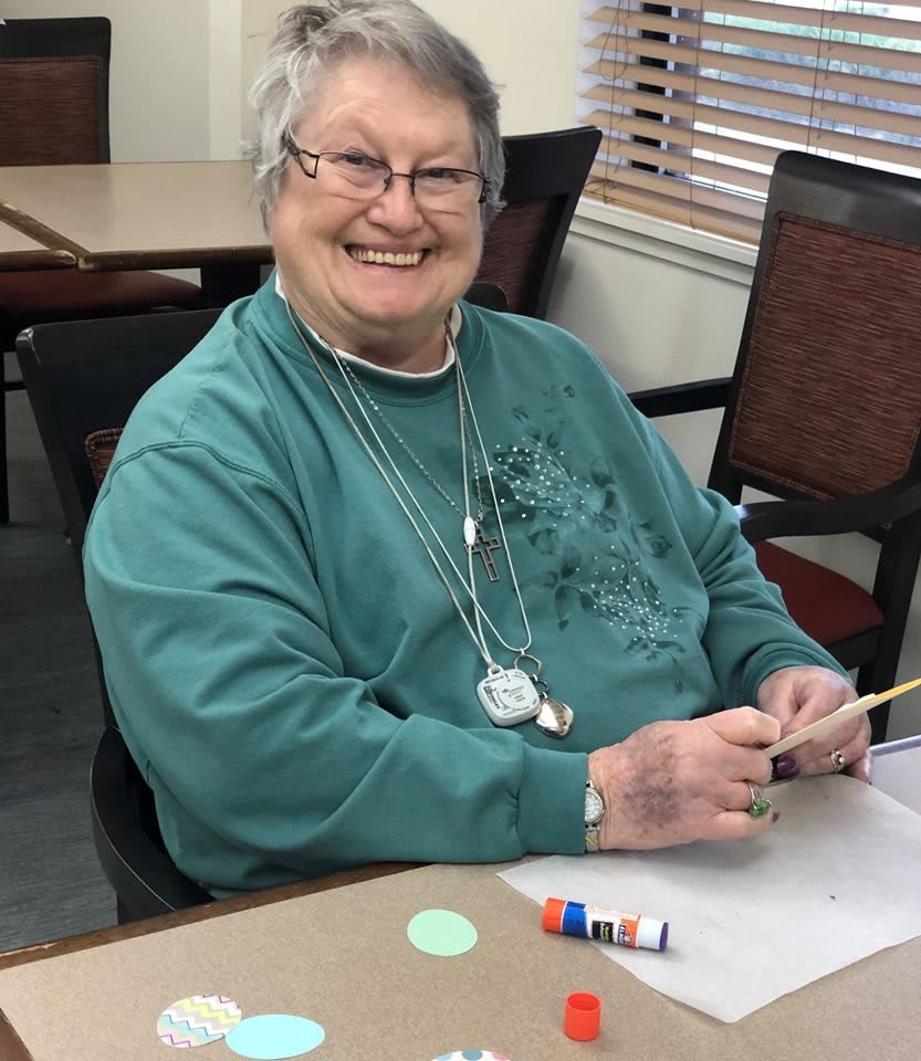 An elderly woman smiles while engaging in a crafting activity at a table.