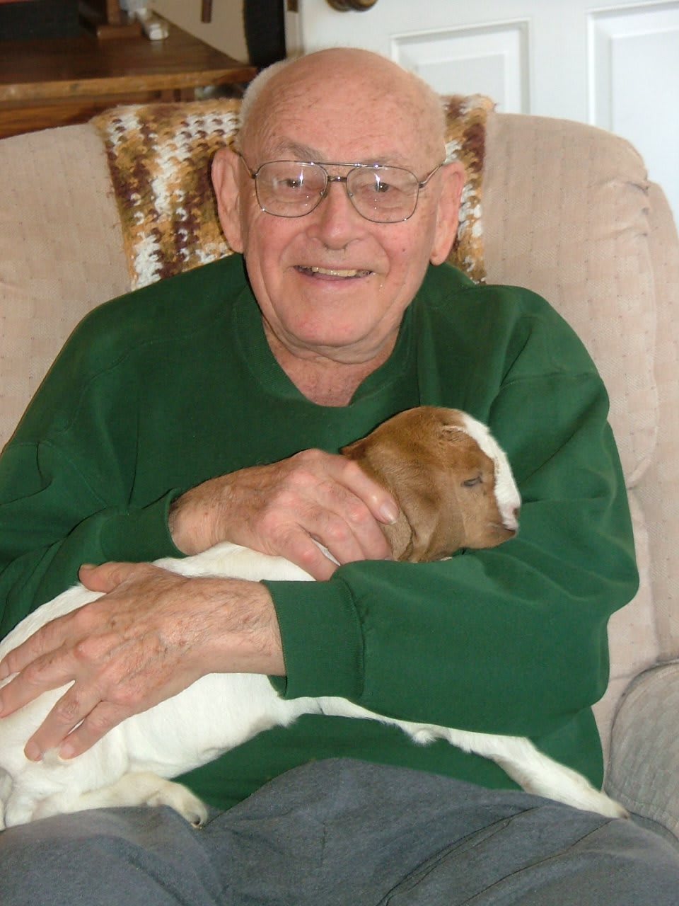 An elderly man wearing glasses and a green sweater smiles while holding a dog indoors.