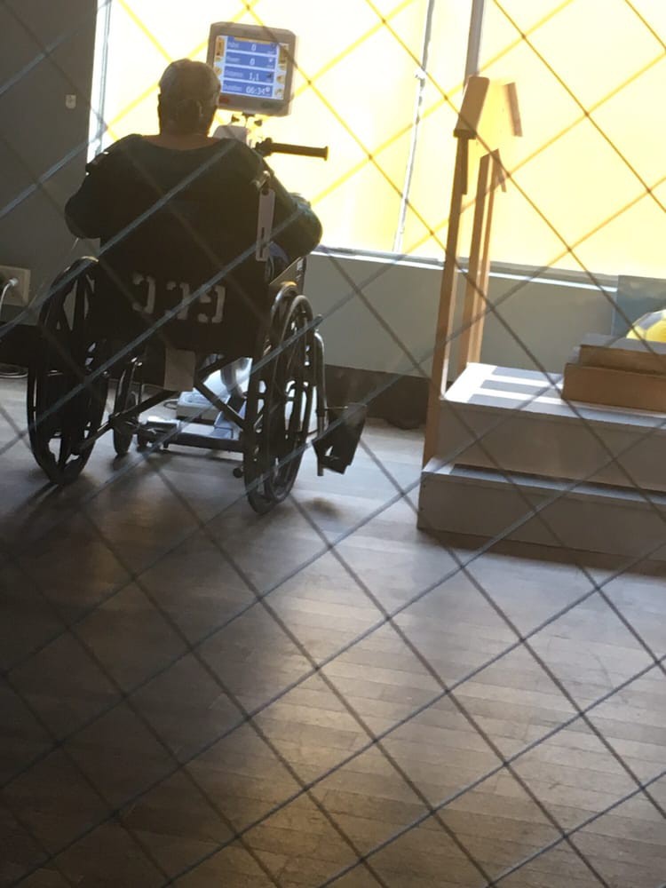 A person sitting in a wheelchair indoors, near a sunlit window and tiled floor.