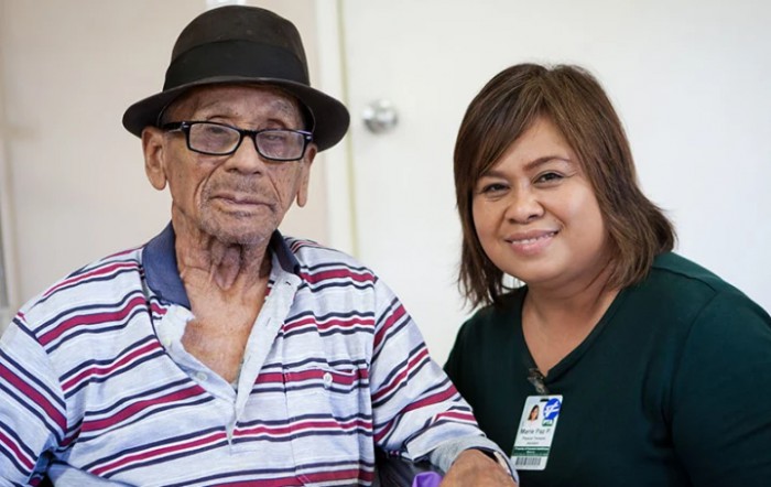An elderly man wearing a hat and glasses sits next to a smiling woman, possibly a caregiver.