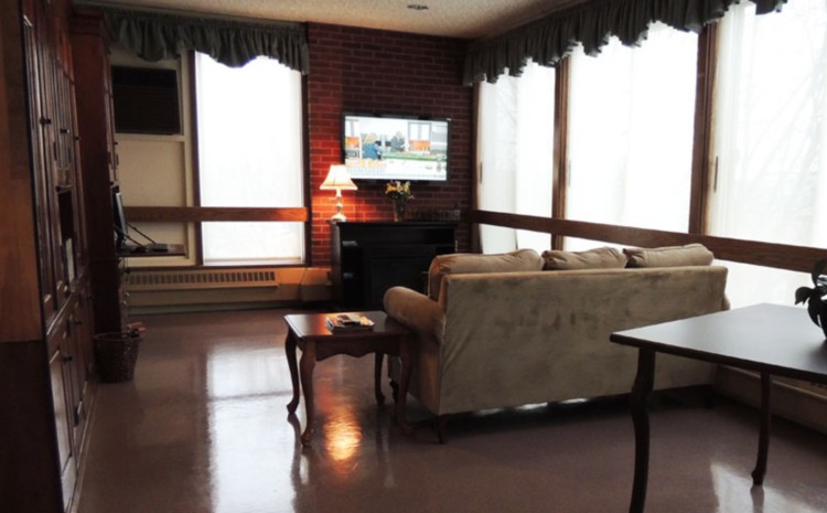 A cozy living room with a sofa and coffee table facing a television mounted on a brick wall, illuminated by a lamp next to large windows.