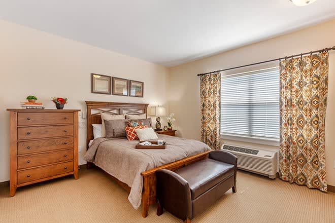 A cozy and elegantly decorated bedroom with a wooden bed frame, matching dresser, and a bench at the foot of the bed. The room is well-lit with natural light coming through a patterned curtain.