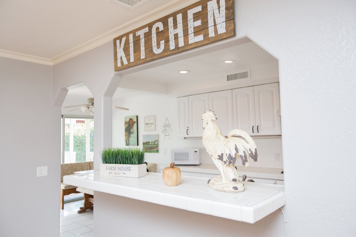 A farmhouse-style kitchen with a white color scheme, featuring a rooster decoration and a wooden 'KITCHEN' sign.