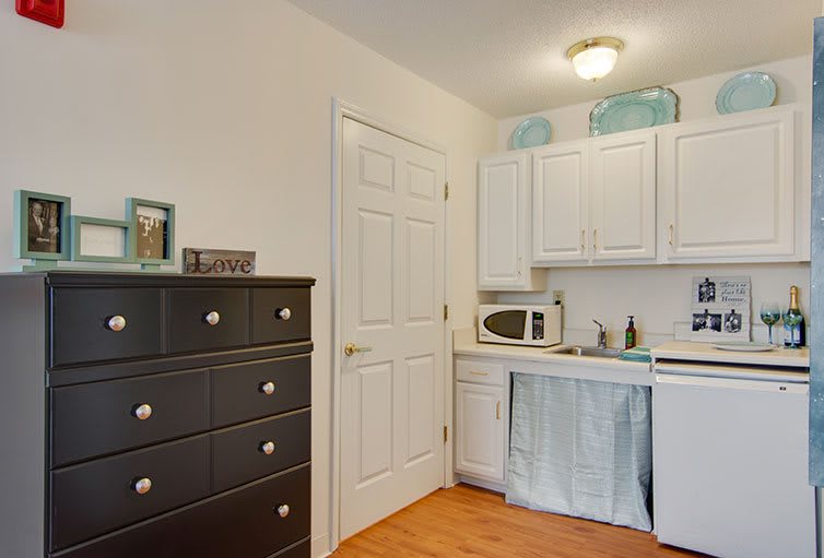 A small kitchen area with white cabinets, a microwave, and a black dresser for additional storage.