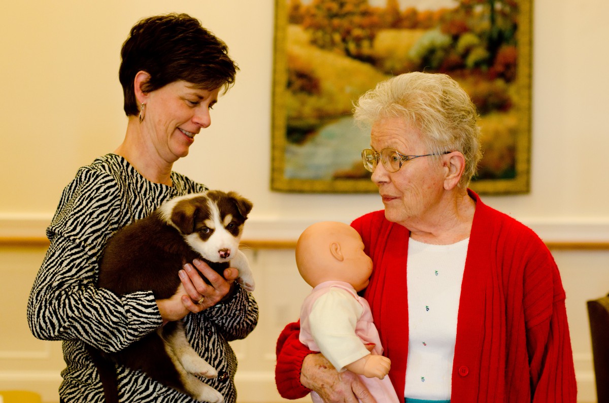 Two women interacting, one holding a puppy and the other a doll.