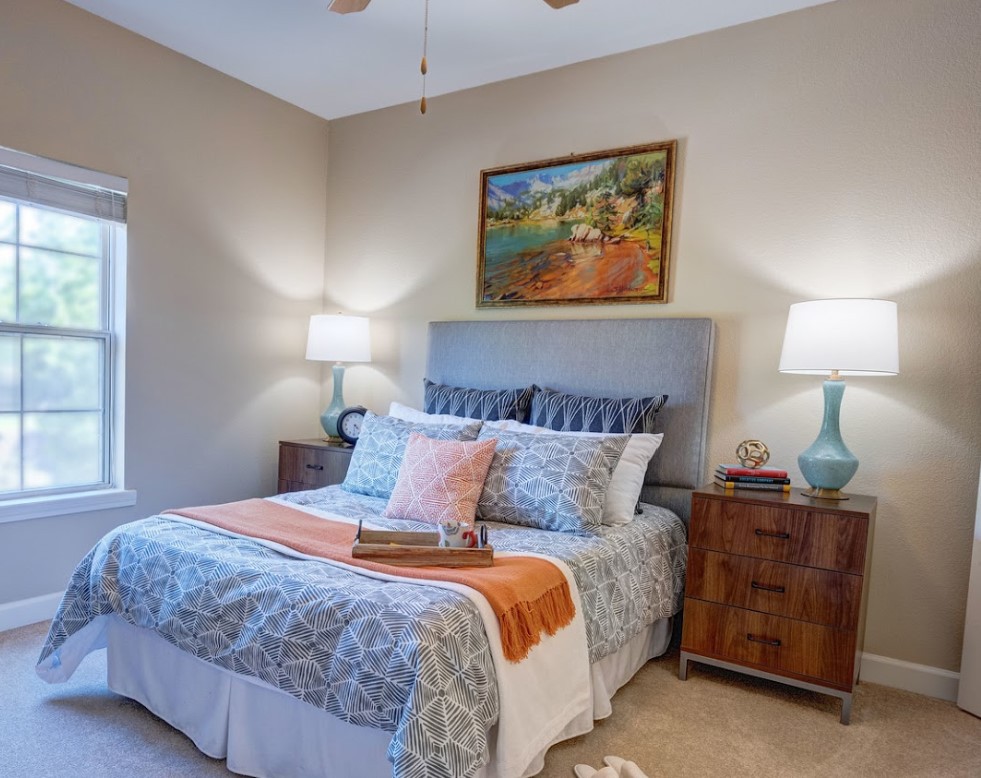 A cozy bedroom with a neatly made bed, featuring colorful bedding and pillows. The room is decorated with lamps on wooden nightstands and a painting above the headboard.