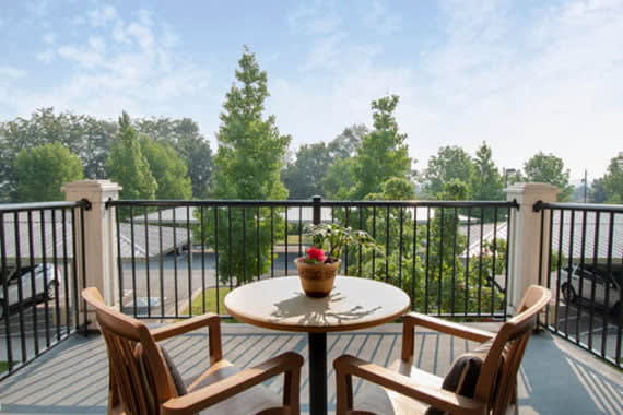 A serene balcony with wooden chairs and a table, overlooking lush green trees.