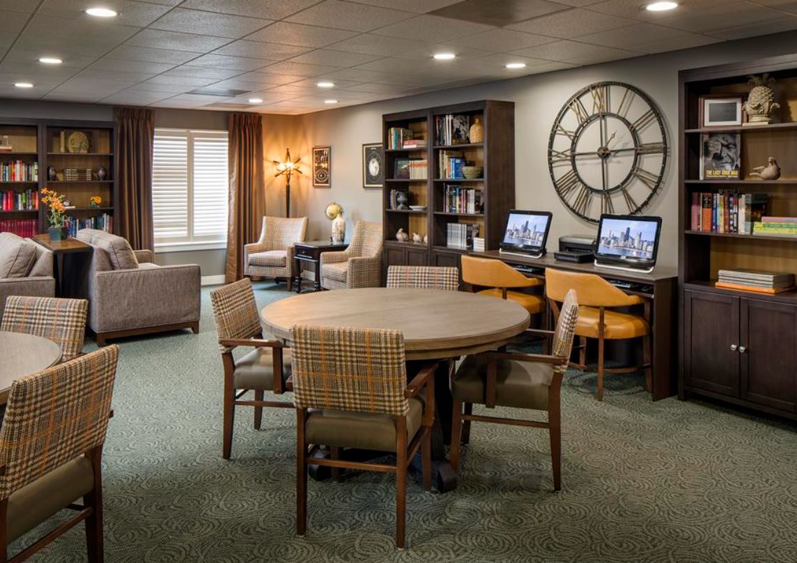 A cozy lounge area with a table and chairs, bookshelves, computers, and a large wall clock.