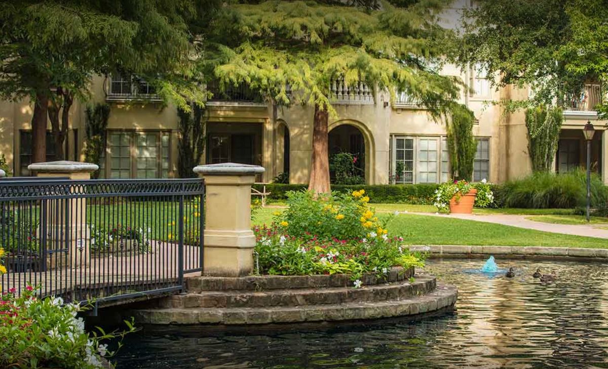 A serene garden landscape featuring a pond, small bridge, and surrounding greenery in front of a building.