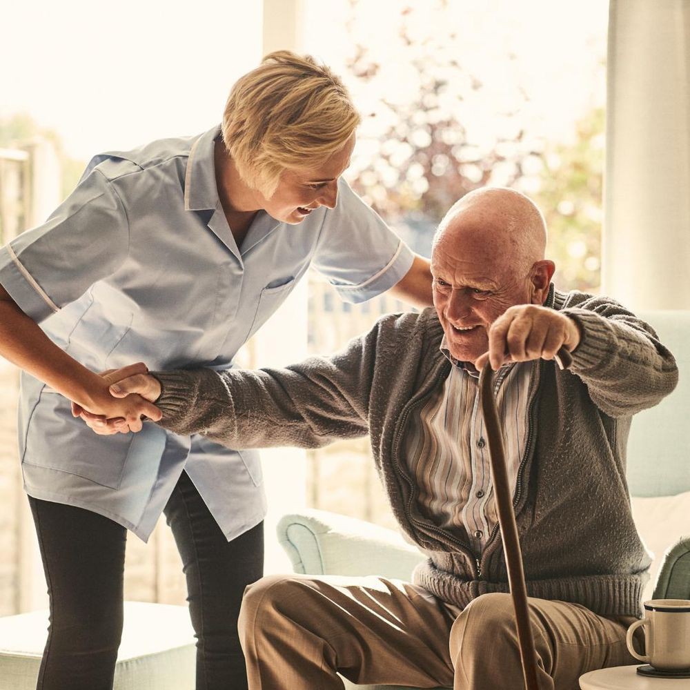 A caregiver assisting an elderly man as he stands up with the help of a cane.