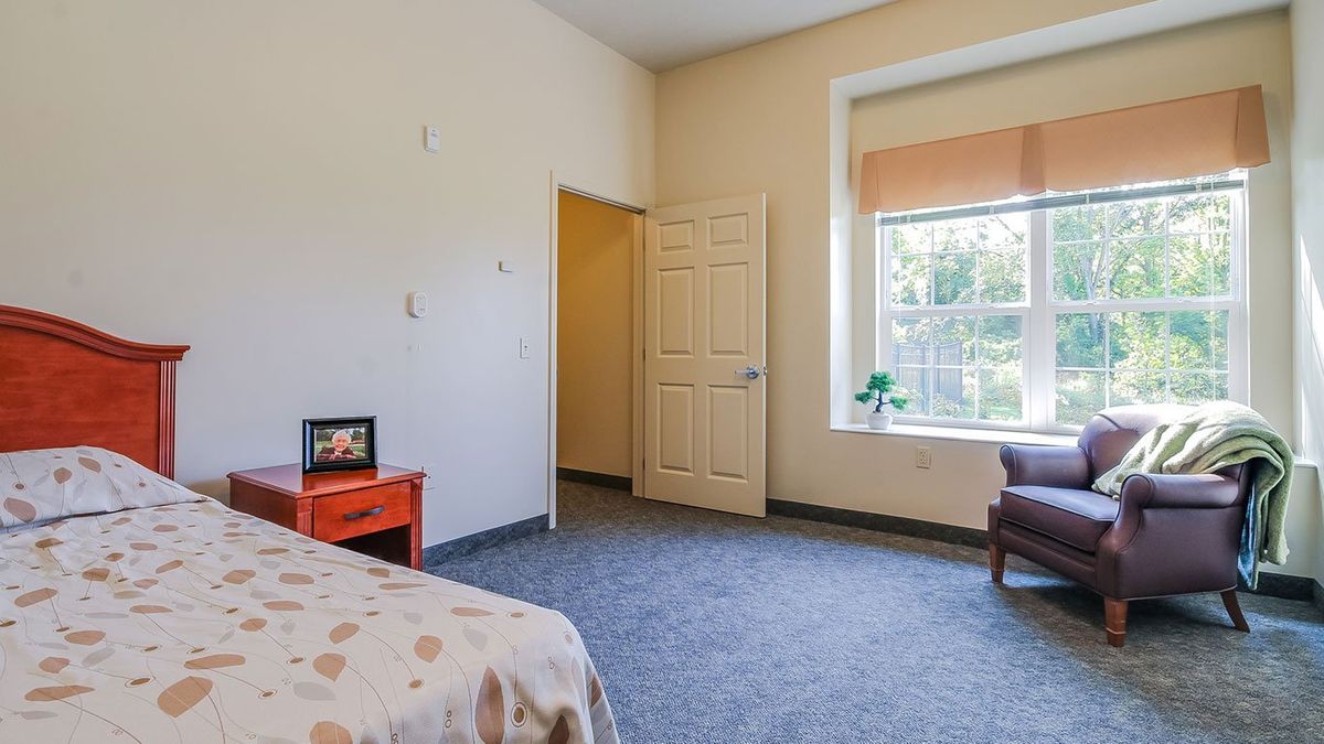 A cozy bedroom with a large window, armchair, and red nightstand.