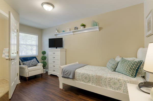 A cozy bedroom featuring a single bed, armchair, and dresser with a TV mounted above. The room is decorated with plants and wall shelves.