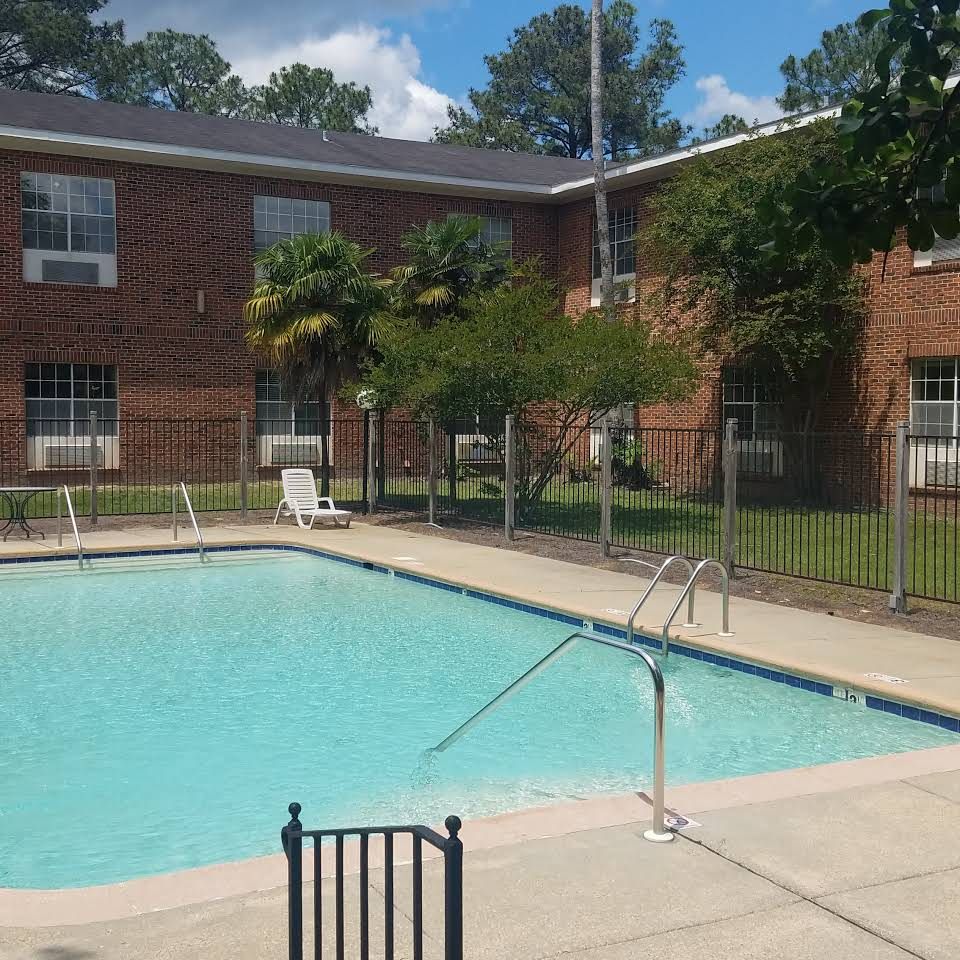A small outdoor swimming pool surrounded by a fence, with a brick building in the background on a sunny day.
