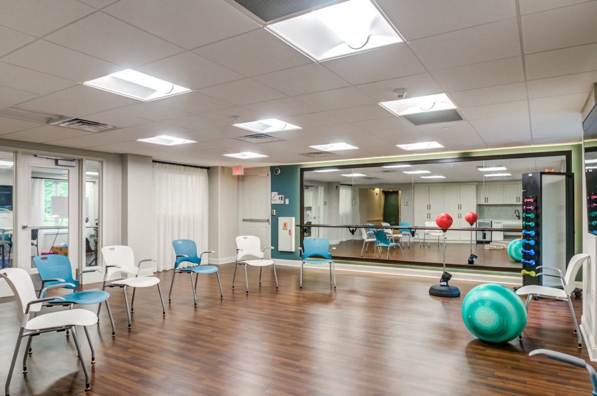 An empty exercise room with chairs, fitness equipment, and mirrors on a wooden floor.