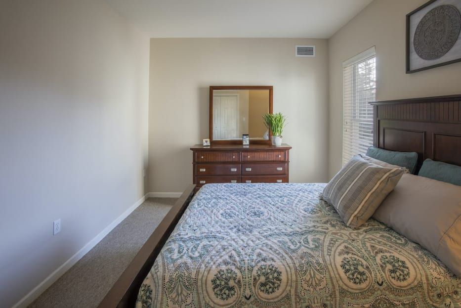 A cozy bedroom with a neatly made bed, a wooden dresser with a mirror, and plenty of natural light.