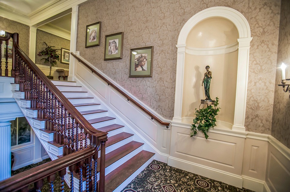 A classic interior with a staircase adorned by framed artwork and a statue in a niche.