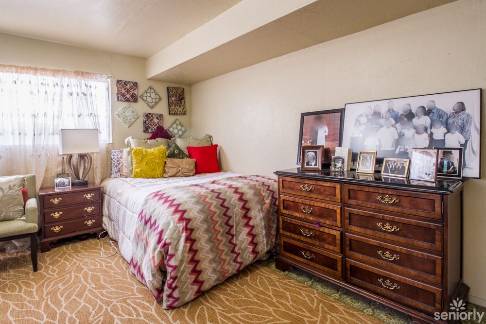 A cozy bedroom with a patterned bedspread, colorful pillows, and a wooden dresser adorned with framed photos.