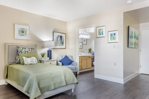 A cozy bedroom with a green bedspread, decorated with tasteful artwork and an adjacent view into the bathroom.