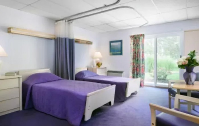A hospital room with twin beds covered in purple bedding and a view of the outside through the window.