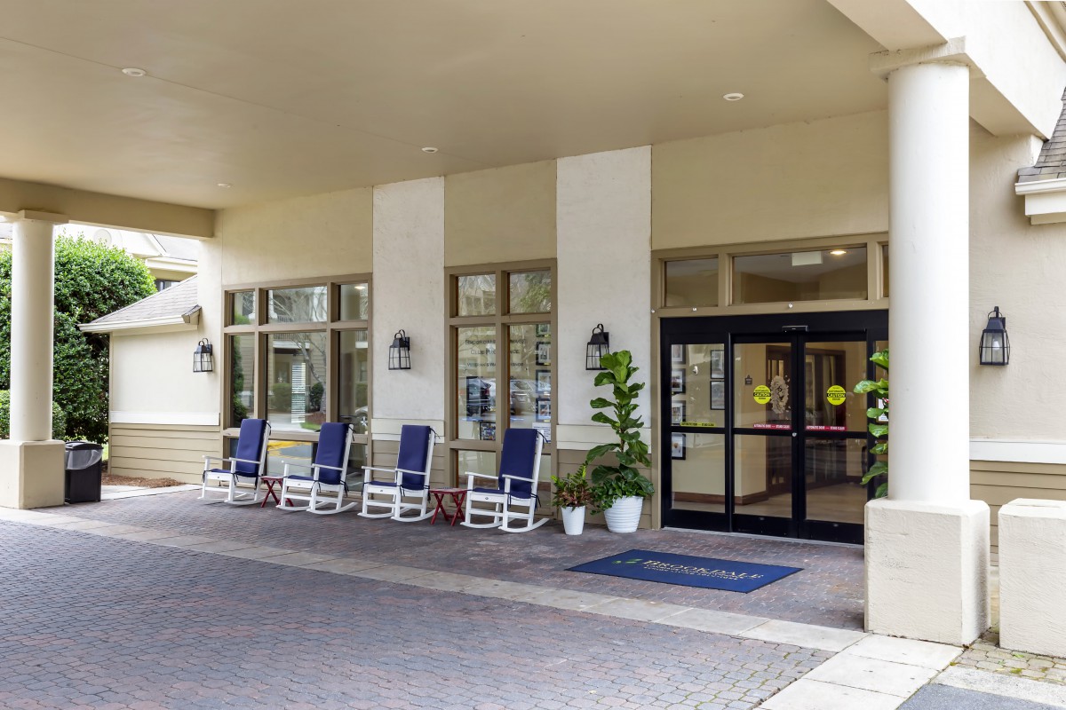 A welcoming building entrance with rocking chairs and potted plants along a covered walkway.