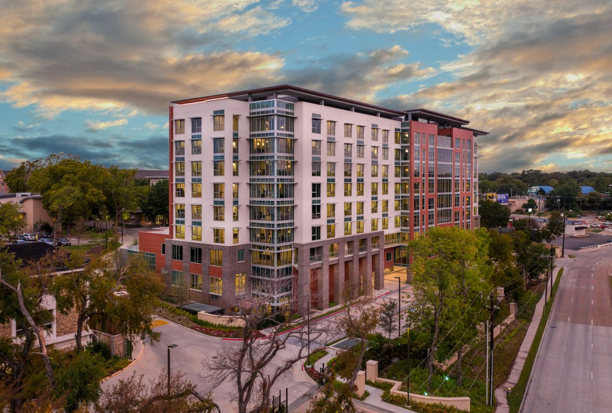 A modern multi-story building surrounded by trees and streets under a cloudy sky.