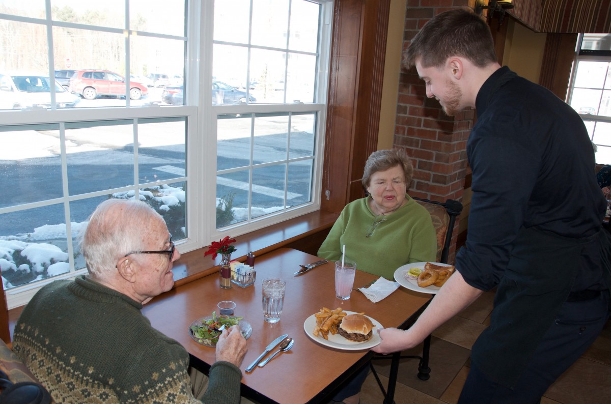 A waiter serves food to two seniors dining at a restaurant.