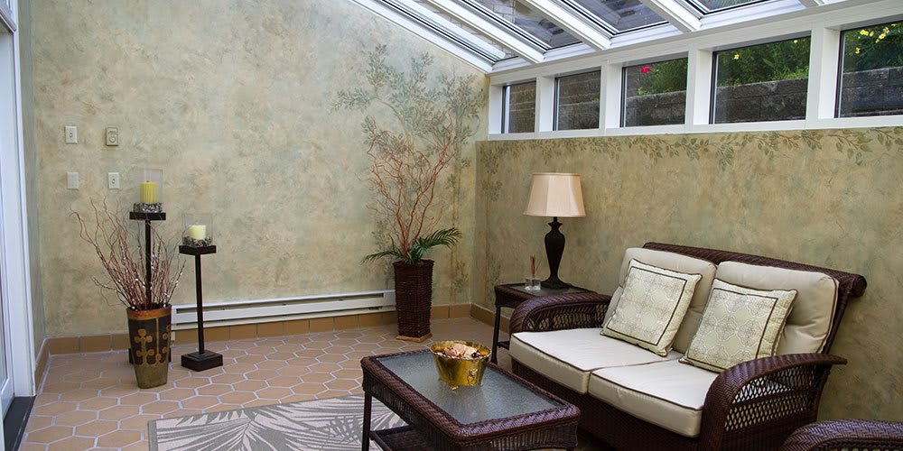 A cozy sunroom with a sofa, table, and lamp, featuring plants and natural lighting.