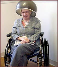 An elderly woman sitting in a wheelchair under a hair dryer at the salon.