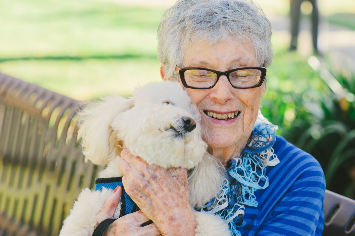 An elderly woman smiling and hugging a fluffy dog outdoors.