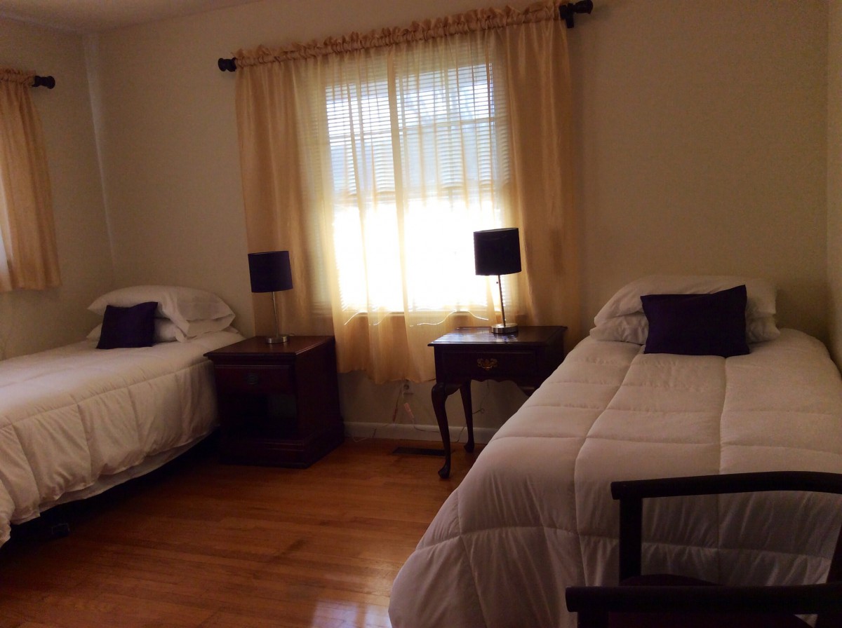 A cozy bedroom featuring twin beds with dark wooden nightstands and lamps, adorned with soft curtains filtering the sunlight through a window.