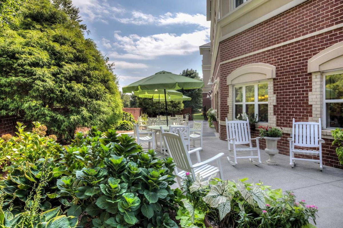 A serene garden patio with white rocking chairs and an umbrella, set against a brick building.
