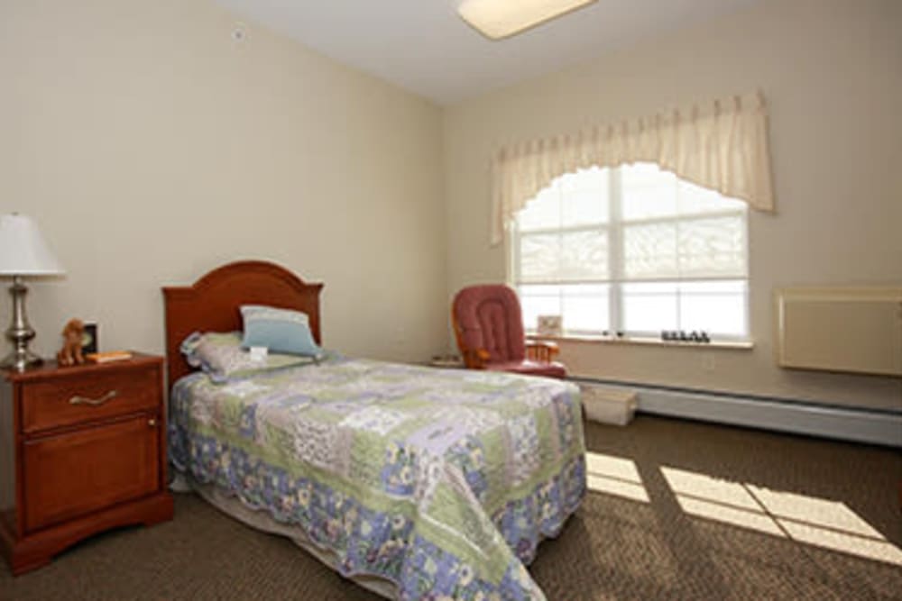 A small, cozy bedroom featuring a single bed with a colorful quilt, next to a large window with curtains. A comfortable chair is placed beside the window.