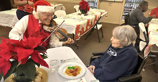 An elderly man in a Santa hat plays the violin for a woman during a festive holiday meal.