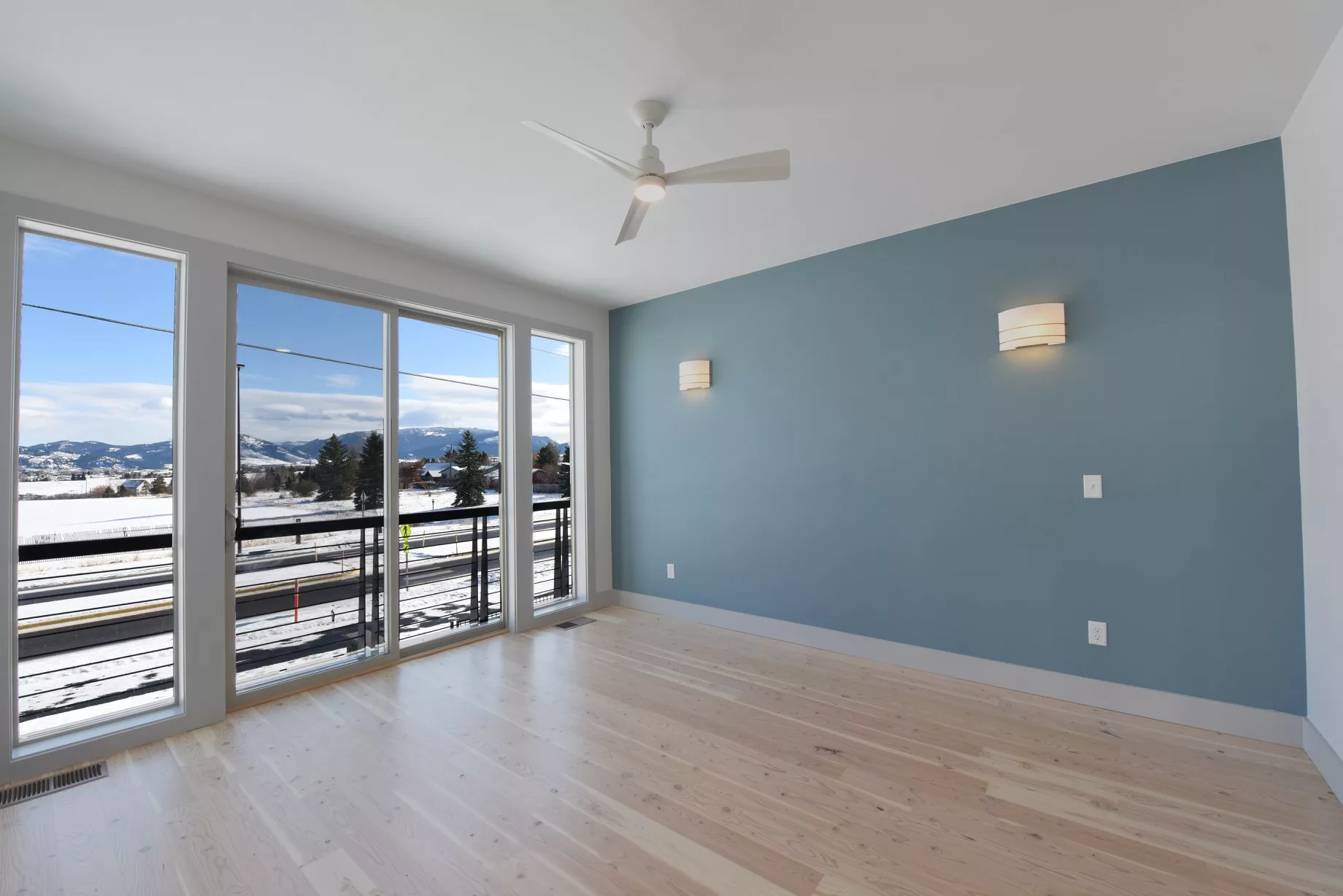 A bright and empty room with wooden flooring, blue accent wall, ceiling fan, and large windows opening to a balcony.