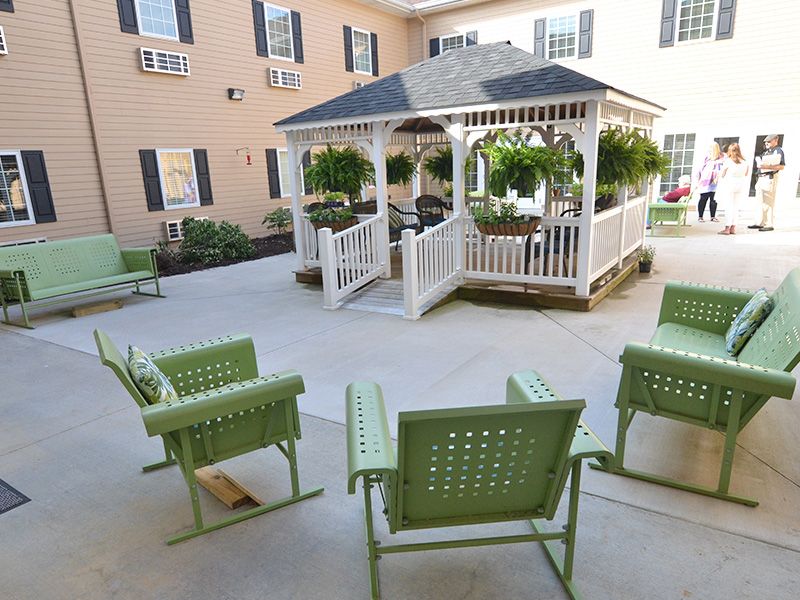 A tranquil courtyard featuring a central gazebo surrounded by green metal chairs and lush hanging plants.