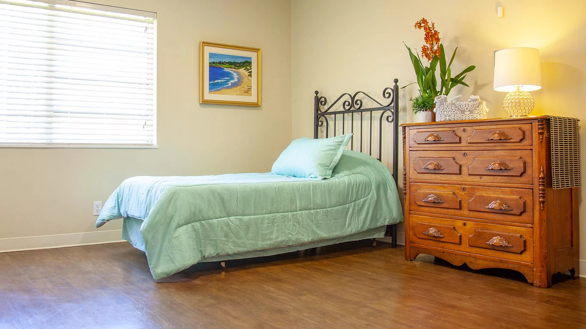 A tidy bedroom with a single bed, green bedding, and a wooden dresser adorned with plants.