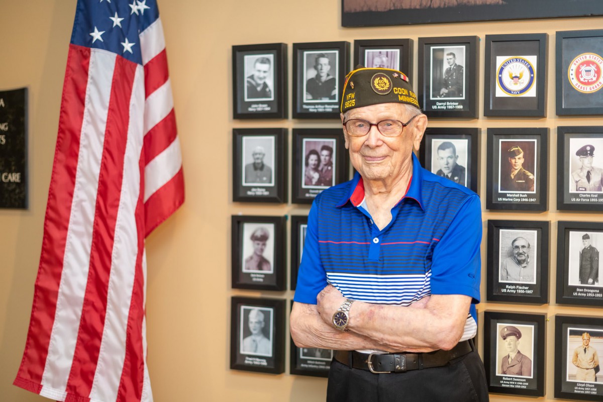 A veteran stands proudly in front of a memorial wall adorned with photos and an American flag.